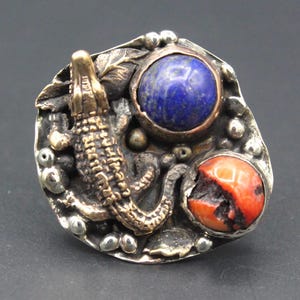 May include: A distinctive silver ring with a bronze alligator motif. The ring includes a large blue lapis lazuli stone and a smaller red coral stone, set within a textured silver setting. The ring has a striking, artistic design.