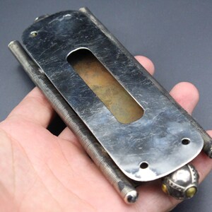 Puede incluir: Objeto met&aacute;lico plateado vintage con forma rectangular y un recorte central. El objeto tiene un mecanismo de resorte a cada lado y una bola decorativa en un extremo. El metal tiene un aspecto desgastado y envejecido.