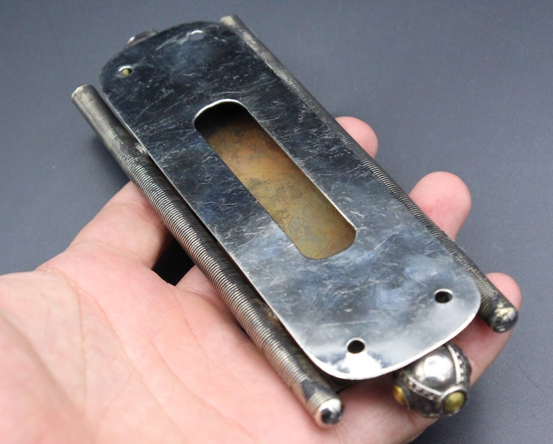 Puede incluir: Objeto met&aacute;lico plateado vintage con forma rectangular y un recorte central. El objeto tiene un mecanismo de resorte a cada lado y una bola decorativa en un extremo. El metal tiene un aspecto desgastado y envejecido.