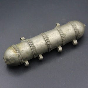 May include: A cylindrical silver-toned metal amulet case featuring a weathered finish, rounded ends, and three decorative braided bands encircling the body. The object includes six small, evenly spaced metal loops attached to the sides.