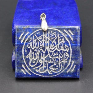 May include: A square, blue lapis lazuli pendant with a silver bail. The pendant features white Arabic calligraphy within a circular design. The stone has a natural, textured appearance. The pendant is likely for a necklace.