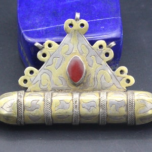 May include: A silver and gold pendant with a red stone in the center. The pendant is in the shape of a triangle with a cylindrical container attached to the bottom.
