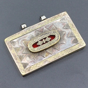 Pode incluir: Um pingente de prata e ouro com um design geométrico vermelho e dourado. O pingente tem formato retangular e é decorado com um padrão de estrela.