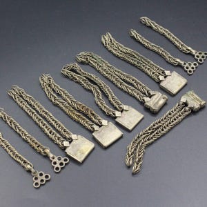 Pode incluir: Uma cole&ccedil;&atilde;o de pe&ccedil;as de joalharia em metal prateado, incluindo pulseiras de corrente tran&ccedil;ada e elementos decorativos. As pe&ccedil;as t&ecirc;m um aspeto vintage, com detalhes quadrados e circulares nas extremidades. Os artigos est&atilde;o dispostos numa superf&iacute;cie cinzenta escura.