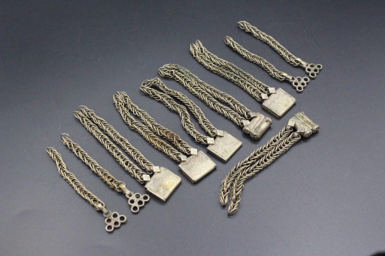 Pode incluir: Uma cole&ccedil;&atilde;o de pe&ccedil;as de joalharia em metal prateado, incluindo pulseiras de corrente tran&ccedil;ada e elementos decorativos. As pe&ccedil;as t&ecirc;m um aspeto vintage, com detalhes quadrados e circulares nas extremidades. Os artigos est&atilde;o dispostos numa superf&iacute;cie cinzenta escura.