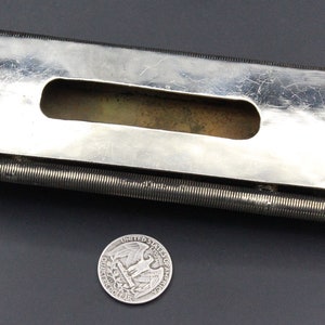 Puede incluir: Un objeto met&aacute;lico plateado con un cuerpo rectangular y un recorte central. Dos varillas enrolladas se extienden desde los lados, y elementos esf&eacute;ricos decorativos est&aacute;n en cada extremo. Una moneda de veinticinco centavos estadounidense est&aacute; al lado del objeto.