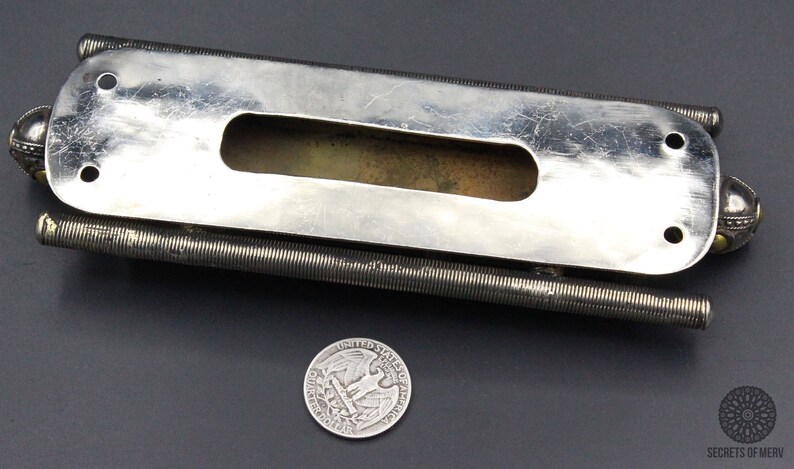 Puede incluir: Un objeto met&aacute;lico plateado con un cuerpo rectangular y un recorte central. Dos varillas enrolladas se extienden desde los lados, y elementos esf&eacute;ricos decorativos est&aacute;n en cada extremo. Una moneda de veinticinco centavos estadounidense est&aacute; al lado del objeto.