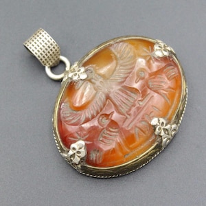 Puede incluir: Colgante ovalado con un diseño tallado que presenta pájaros y otras figuras en tonos naranja y marrón. El colgante tiene un marco de metal plateado con detalles florales y una fianza texturizada para una cadena.