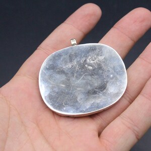 May include: A silver pendant with a smooth, rounded surface. The pendant has a loop at the top for attaching a chain.