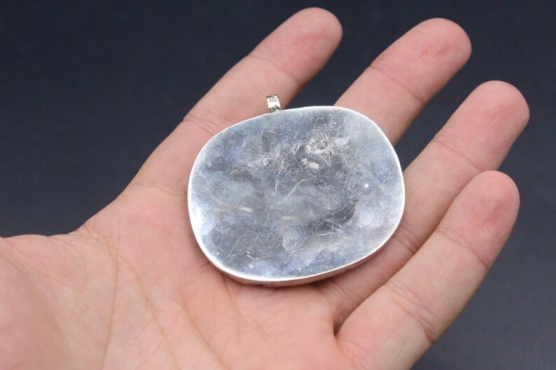 May include: A silver pendant with a smooth, rounded surface. The pendant has a loop at the top for attaching a chain.