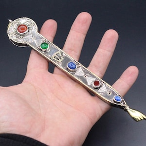 Può includere: Un oggetto decorativo argentato e ornato con pietre preziose incastonate rosse, verdi e blu. Il pezzo ha una forma a chiave con un'estremità appuntita e una piccola mano dorata che indica. L'oggetto è tenuto in una mano su sfondo scuro.