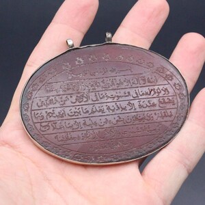 Könnte beinhalten: Ovaler, rotbrauner Anhänger mit arabischer Schrift, eingefasst in einen silberfarbenen Metallrahmen. Der Anhänger hat oben zwei kleine Schlaufen. Der Text ist in mehreren Zeilen eingraviert. Der Anhänger wird in einer Hand gehalten.