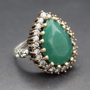 May include: A silver ring with a large, pear-shaped, faceted green gemstone. The gemstone is surrounded by a halo of small, clear gemstones and gold-colored accents. The ring has a detailed, ornate design.