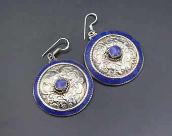 May include: A pair of silver-toned earrings with a circular design. The earrings feature a blue border and a central blue stone. The center of the earrings have a floral pattern. The earrings have a hook closure.