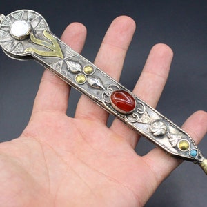 Peut inclure: Un objet décoratif orné en argent et or avec des pierres précieuses rouges et bleues. L'objet a une extrémité pointue et un sommet arrondi avec une petite pierre blanche.