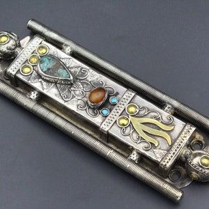 Puede incluir: Una caja de metal plateado adornada con piedras preciosas turquesas y naranjas, y detalles dorados. La caja tiene forma rectangular con dos varillas met&aacute;licas en espiral paralelas a cada lado.