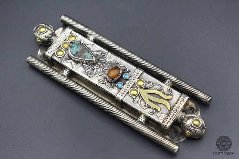 Puede incluir: Una caja de metal plateado adornada con piedras preciosas turquesas y naranjas, y detalles dorados. La caja tiene forma rectangular con dos varillas met&aacute;licas en espiral paralelas a cada lado.