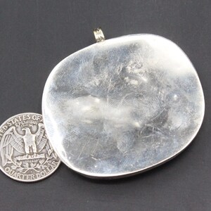 May include: A silver pendant with a smooth, rounded surface. The pendant has a loop at the top for attaching a chain.
