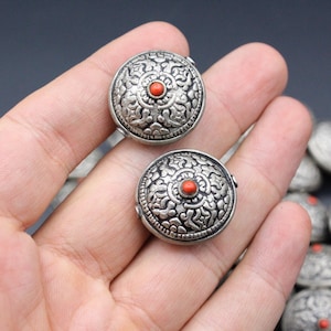 Könnte beinhalten: Nahaufnahme einer Hand, die zwei runde Knöpfe aus Silber und Koralle mit filigranen Designs hält. Die Knöpfe sind von einem Stapel ähnlicher Knöpfe umgeben.