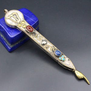 Può includere: Un pendente in argento e oro ornato con pietre preziose intarsiate, tra cui rosse, blu e turchesi. Il pendente presenta un design stilizzato con una mano che indica verso il basso. Poggia su un blocco blu rettangolare.