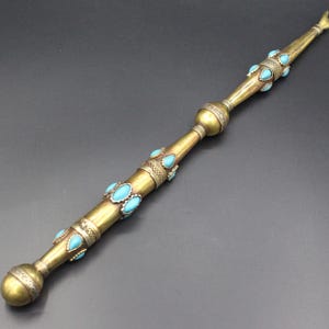 Könnte beinhalten: Ein langer, verzierter Zauberstab mit Messing-Finish und türkisfarbenen Steinakzenten. Der Zauberstab hat einen abgerundeten Griff, dekorative Bänder und eine spitze Spitze. Die türkisfarbenen Steine sind tropfenförmig und entlang des Schafts angeordnet.