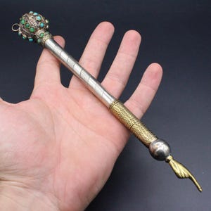Può includere: Un puntatore in metallo con una parte superiore sferica decorata con piccole pietre turchesi e verdi, un'asta argentata con un motivo inciso in diagonale, una sezione di presa dorata testurizzata e una mano color ottone con l'indice teso.