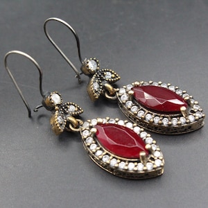 May include: A pair of drop earrings featuring a central marquise-cut deep red stone surrounded by a halo of small clear white crystals. The setting is an antiqued bronze-toned metal with a floral-inspired top piece and dark metal hooks.