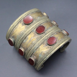 May include: A wide cuff bracelet made of tarnished brass featuring three horizontal bands with intricate etched leaf patterns. The bracelet is decorated with six oval-shaped, smooth red stones set in metal bezels along the edges and center.
