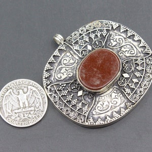May include: A silver pendant with an intricate design and a red stone in the center. The pendant is about the size of a US quarter.