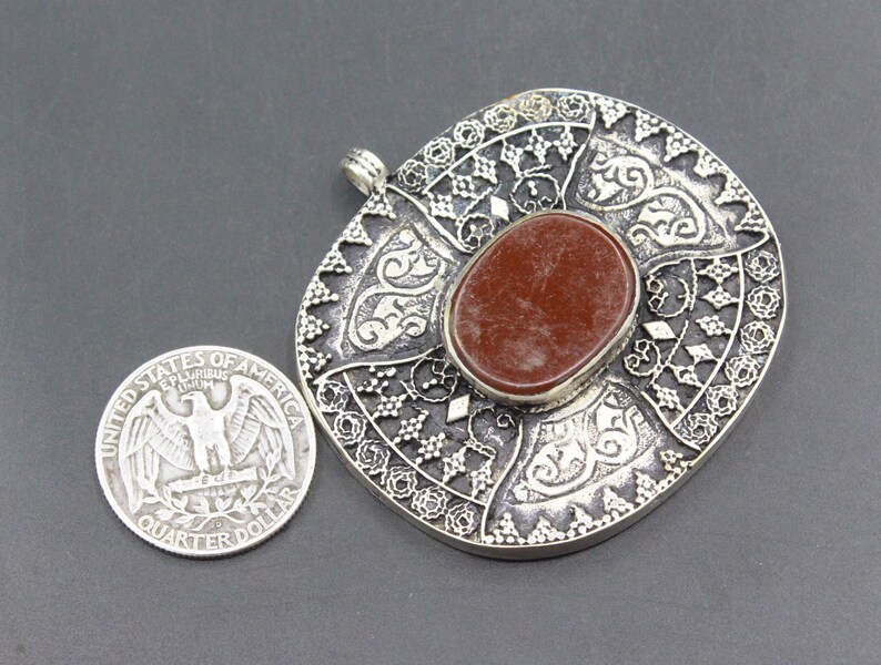 May include: A silver pendant with an intricate design and a red stone in the center. The pendant is about the size of a US quarter.