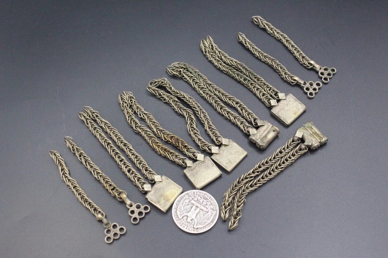 Pode incluir: Uma cole&ccedil;&atilde;o de pe&ccedil;as de corrente de metal prateado antigas, algumas com elementos decorativos quadrados ou circulares. As correntes est&atilde;o dispostas sobre uma superf&iacute;cie cinza escuro, com uma moeda vis&iacute;vel para escala.
