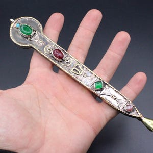 Può includere: Un oggetto in metallo argentato ornato con pietre preziose verdi, rosse e rosa. Il pezzo allungato presenta disegni intricati e un piccolo dettaglio a forma di mano all'estremità. Una pietra turchese è incastonata nella parte superiore.