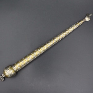 Può includere: Un oggetto metallico lungo, sottile e affusolato con una base arrotondata e bulbosa e una punta acuminata. La superficie presenta un motivo alternato di forme geometriche color argento e oro, inclusi triangoli e rombi lungo tutta la lunghezza.