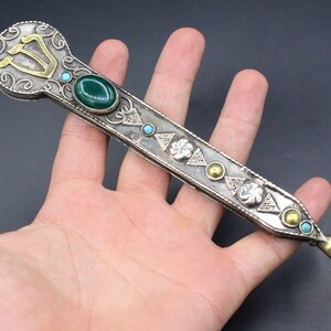 May include: A silver and gold pointer with a green stone and turquoise accents. The pointer has a decorative design and a hand-shaped tip.