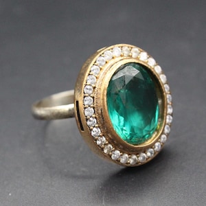 May include: An oval-cut emerald ring with a halo of small, clear gemstones set in a gold-colored band. The ring's band is silver-colored. The emerald has a deep green hue, and the ring is set against a dark background.