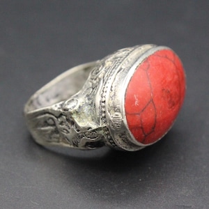 Può includere: Anello color argento con una pietra rossa brillante di forma ovale. La fascia dell'anello presenta dettagli intricati in rilievo. La pietra è incastonata in una lunetta d'argento, su sfondo scuro.