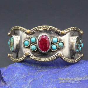 May include: Silver cuff bracelet with a central oval ruby and turquoise accents. The bracelet has a decorative gold-colored trim and is set on a blue surface. A unique piece of jewelry.