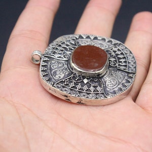 May include: A silver pendant with intricate designs and a red stone in the center. The pendant is round and has a loop at the top for attaching a chain.