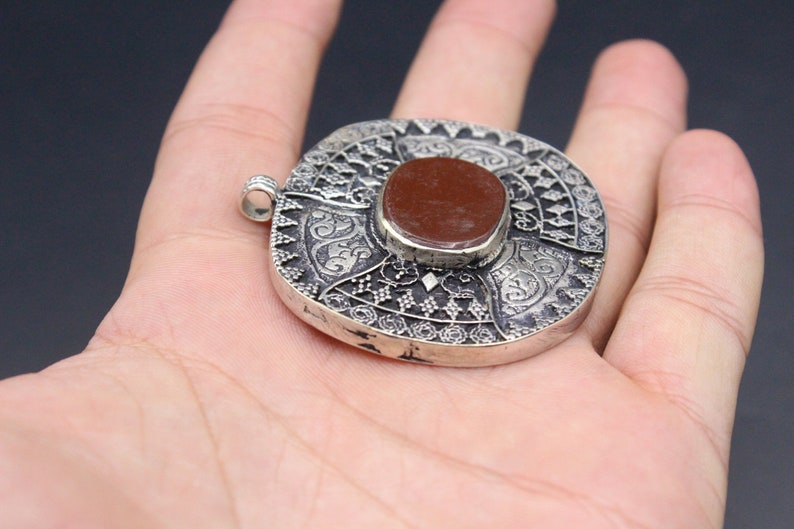 May include: A silver pendant with intricate designs and a red stone in the center. The pendant is round and has a loop at the top for attaching a chain.