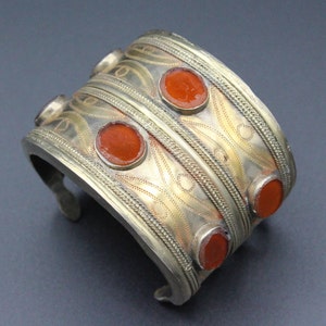May include: Silver-toned cuff bracelet with six round, orange-red stones. The bracelet features intricate etched designs and braided edges. A vintage-style accessory.