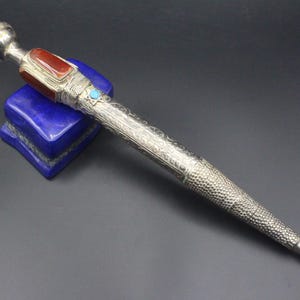 Könnte beinhalten: Ein silberner und roter, mit Edelsteinen besetzter Stift mit aufwendigen Gravuren. Der Stift hat eine runde, silberne Spitze und eine spitze Spitze. Er liegt auf einem blauen Steinsockel.