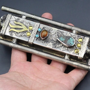 Puede incluir: Objeto de metal plateado adornado con detalles dorados y piedras de color turquesa y &aacute;mbar. La pieza rectangular tiene dos varillas met&aacute;licas enrolladas que se extienden desde los lados. Pieza decorativa con detalles intrincados.