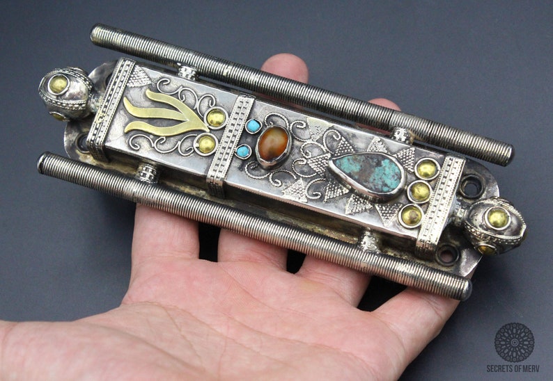 Puede incluir: Objeto de metal plateado adornado con detalles dorados y piedras de color turquesa y &aacute;mbar. La pieza rectangular tiene dos varillas met&aacute;licas enrolladas que se extienden desde los lados. Pieza decorativa con detalles intrincados.