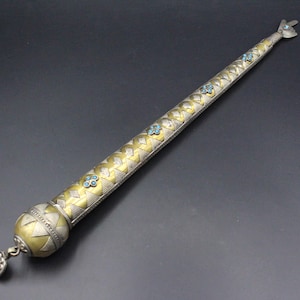 Può includere: Un lungo oggetto metallico affusolato con finitura oro e argento, dotato di una base sferica con un piccolo ciondolo pendente. La superficie è decorata con un motivo a rombi ripetuto e piccole pietre blu incastonate lungo la lunghezza.