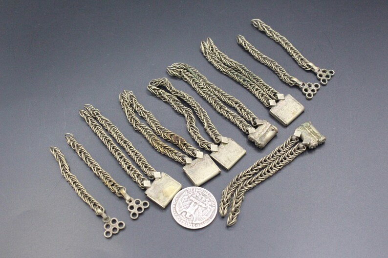 Pode incluir: Uma variedade de pulseiras antigas de corrente prateada com fechos quadrados e extremidades decorativas. As pulseiras est&atilde;o dispostas sobre uma superf&iacute;cie escura, com uma moeda de um quarto de d&oacute;lar americano para escala.