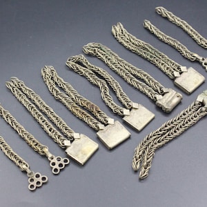 Pode incluir: Uma cole&ccedil;&atilde;o de pe&ccedil;as de joalharia em metal prateado. As pe&ccedil;as apresentam um design de corrente tran&ccedil;ada, algumas com fechos quadrados e outras com extremidades decorativas vazadas. Os artigos est&atilde;o dispostos sobre uma superf&iacute;cie cinza escuro.