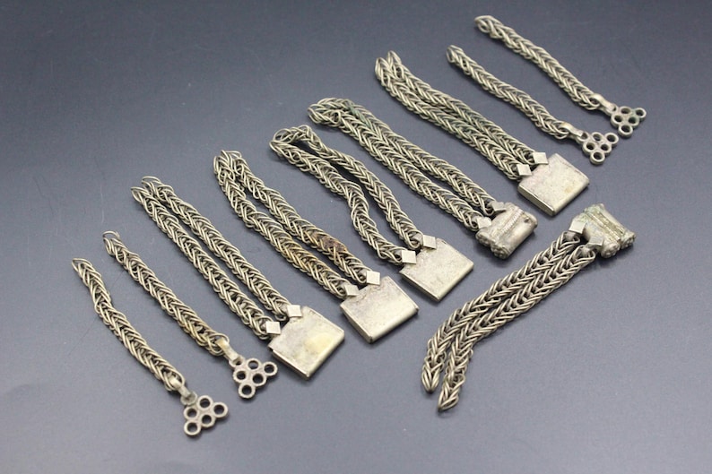 Pode incluir: Uma cole&ccedil;&atilde;o de pe&ccedil;as de joalharia em metal prateado. As pe&ccedil;as apresentam um design de corrente tran&ccedil;ada, algumas com fechos quadrados e outras com extremidades decorativas vazadas. Os artigos est&atilde;o dispostos sobre uma superf&iacute;cie cinza escuro.