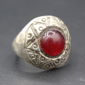 May include: A silver ring with a large, round, red gemstone at the center. The ring's band is wide and features intricate etched patterns around the gemstone. The ring has a vintage or antique appearance.
