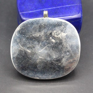 May include: Silver pendant with a smooth, oval-shaped front. The pendant has a loop at the top for attaching a chain.