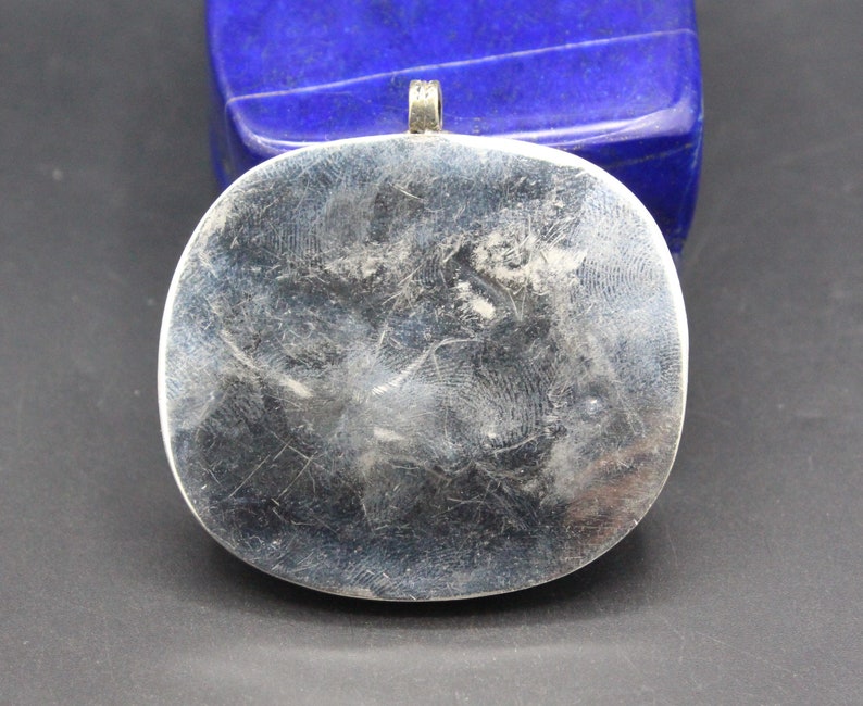 May include: Silver pendant with a smooth, oval-shaped front. The pendant has a loop at the top for attaching a chain.